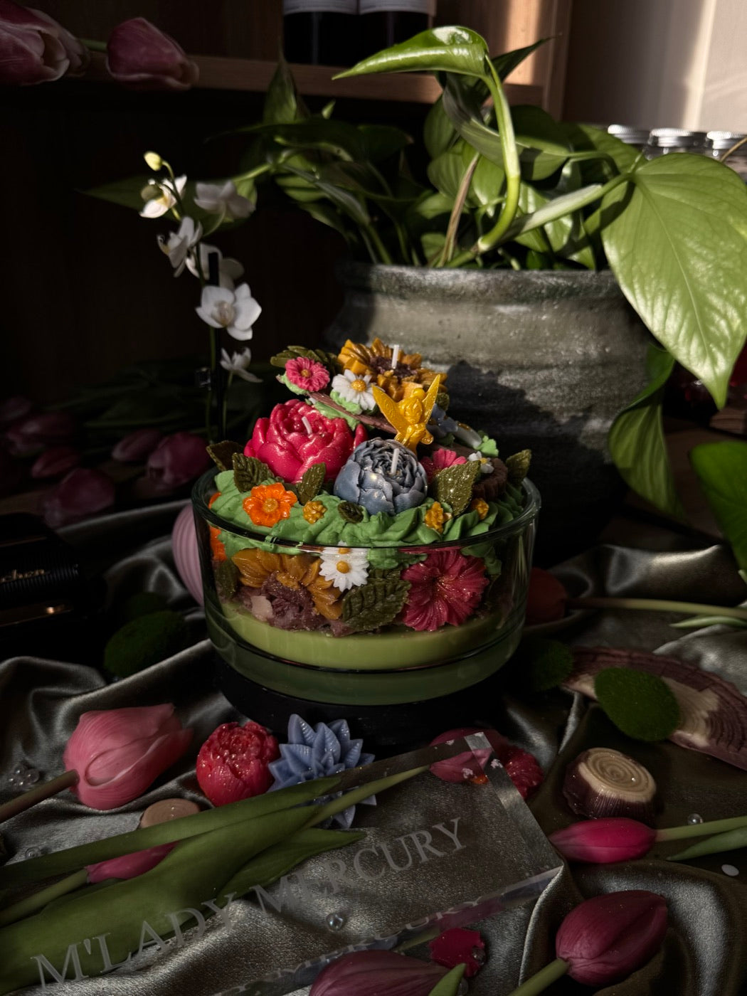 A decorative candle in a glass bowl, topped with wax roses, sunflowers and green leaves